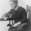 Seated woman in dark coloured dress with beading, white gloves and holding a fan and fresh cut roses