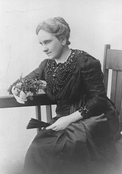 Seated woman in dark coloured dress with beading, white gloves and holding a fan and fresh cut roses