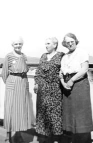 Three women stand on the deck of a ship with hills in the background