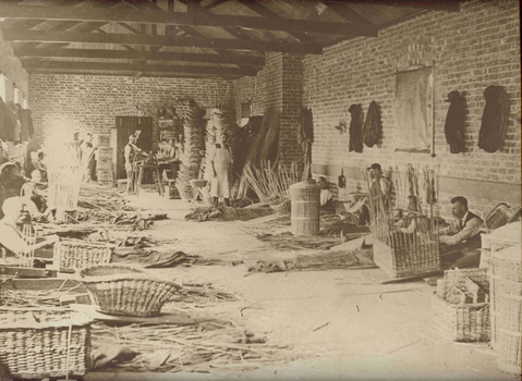 Men sitting on hessian making cane baskets inside a workshop