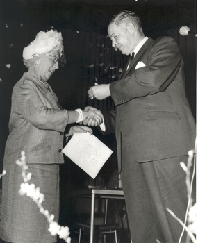 Mrs Tutton shaking hands with a suited man and receiving something, possibly a badge