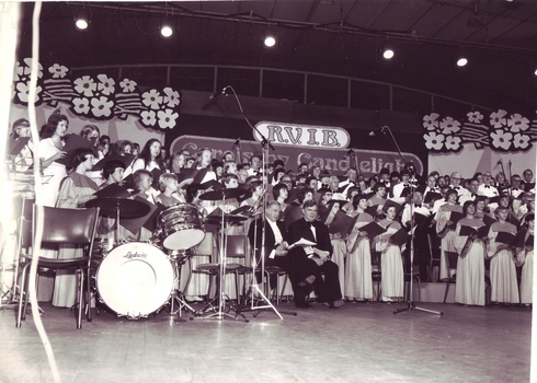 Two men in black suits sit amongst standing choristers on the Carols stage