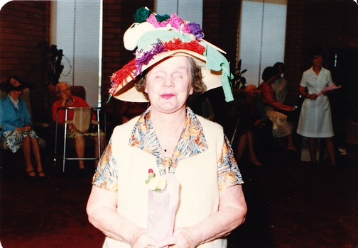Woman wearing a hat with skeins of yarn and knitting needles on top, holding flowers