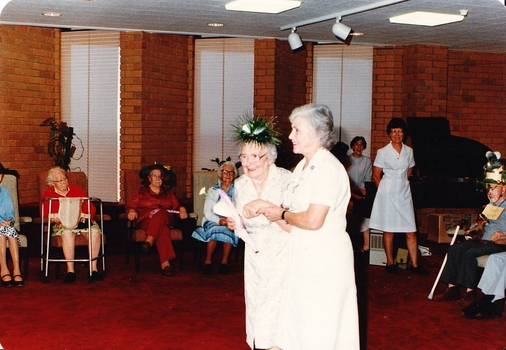 A nursing sister helps an older woman wearing a green spikey hat with tinsel across the floor