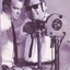 One man with ear muffs holding a rifle attached to a frame while another man looks on