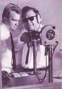 One man with ear muffs holding a rifle attached to a frame while another man looks on