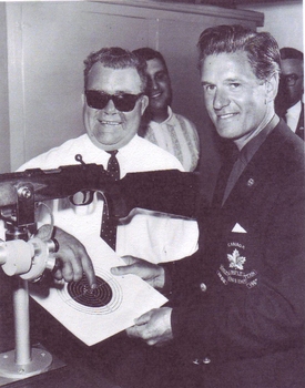 Smiling man points to the bulls eye hit on a target which is held by a man wearing a Canada Rifle club blazer