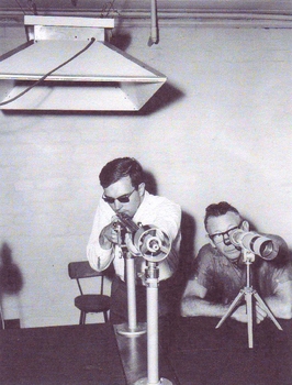 A man holds a rifle in a frame whilst another looks at the target through a telescope