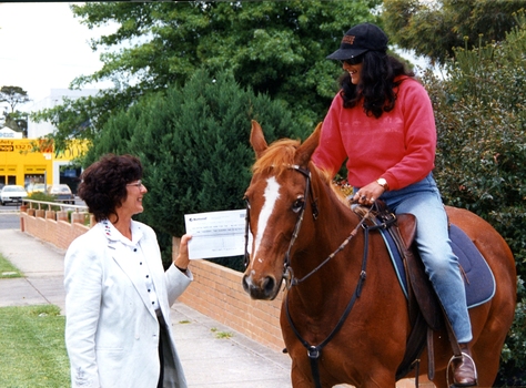 Woman takes an oversized cheque from another woman on horseback