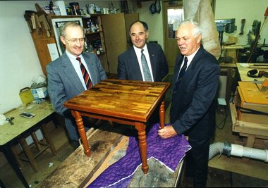 Two men hold a side table whilst a third man looks on 