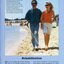 A man holding a white cane walks along a beach whilst holding the arm of a female.