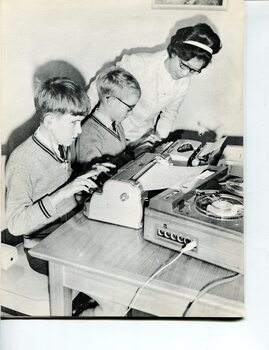 Two children type - one on a Perkins, the other on a typewriter - as a teacher looks on