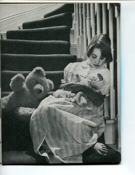 Girl in nightdress sits at the base of stairs with a teddy bear and cuddling a doll