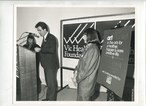 Paul Korsten speaking behind a lectern at the Braille Book of the Year awards 1990 flanked by Fran Awcock and Wilma Bedford