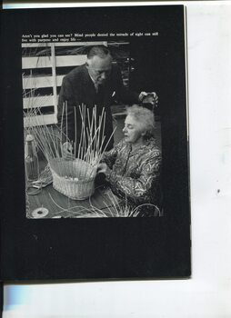 Woman weaving a cane basket whilst a man stands beside her touching the cane stalks