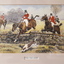 Men dressed in red coats and white shirts and jodhpurs jumping horses over fences following a hunting dog
