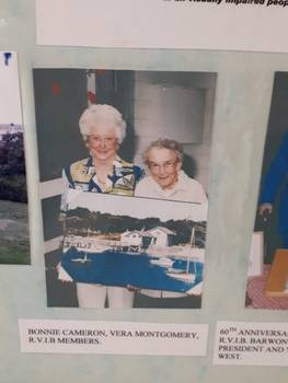Two women holding a painting of the boatshed at Barwon Heads