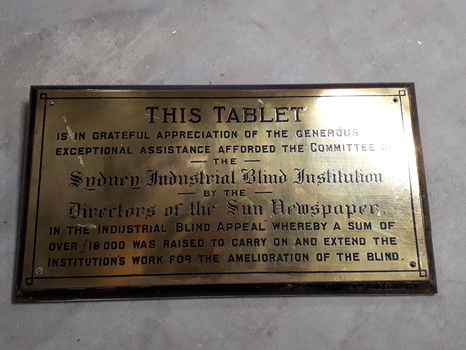 Gold coloured metal plaque with black writing mounted on wood.