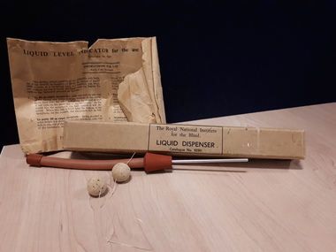 Brown paper packet instructions with box labelled 'Liquid dispenser', red rubber tube, 2 cork balls on string and metal prong
