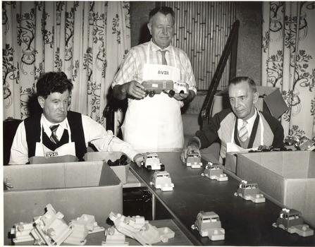 Two seated men put truck components on a production line, with a third man standing holding a truck and trailer.