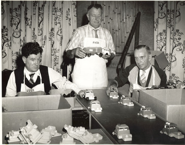 Two seated men put truck components on a production line, with a third man standing holding a truck and trailer.