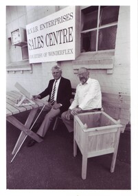 Two men sit on a wooden bench underneath a sign that reads RVIB Enterprises Sales Centre courtesy of Wonderflex