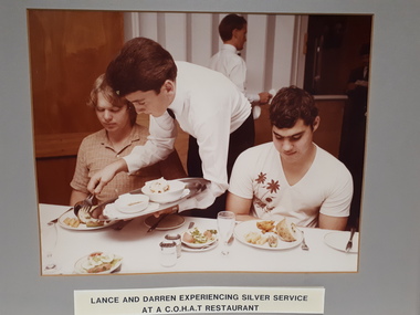 Two boys sit at a table with food on their plates, as the waiter finishes placing potatoes one of the plates.