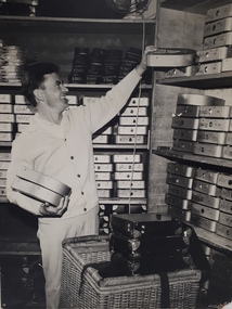 Man holding audio cartridges and lifting another down from a shelf.  A wicker basket with cartridge cases sits before him.