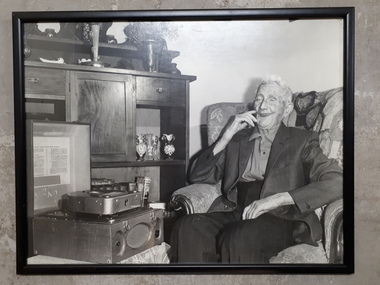 Elderly man sitting next to sideboard, smiling as he listens to a Clarke & Smith cartridge Talking Book Machine