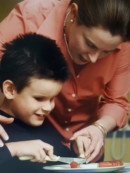 A woman assists a boy cut his hot dog on a plate. 