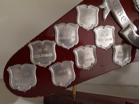Wooden boomerang with various burnished shields, banner and circle with wreath, and golden man holding shield with bowler etched