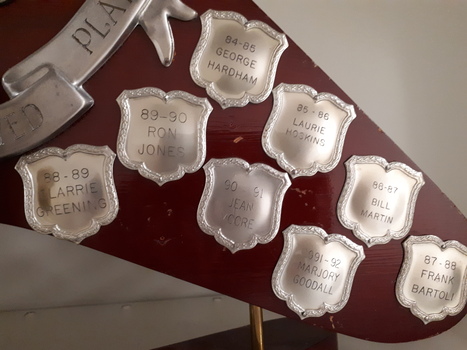 Wooden boomerang with various burnished shields, banner and circle with wreath, and golden man holding shield with bowler etched