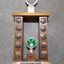Wooden rectangle topped with a plastic wreath and tarnished silver plates inscribed with winner names