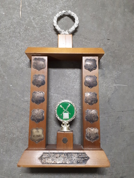 Wooden rectangle topped with a plastic wreath and tarnished silver plates inscribed with winner names