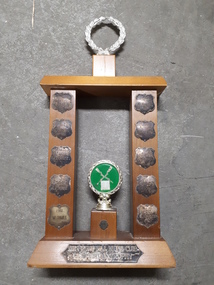 Wooden rectangle topped with a plastic wreath and tarnished silver plates inscribed with winner names