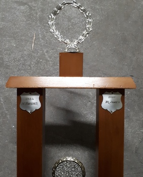 Wooden rectangle topped with a plastic wreath and tarnished silver plates inscribed with winner names