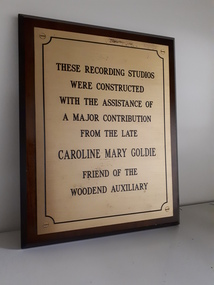 Gold coloured plate with black writing on wooden rectangle