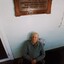 Elderly man sits beneath wooden plaque on verandah.