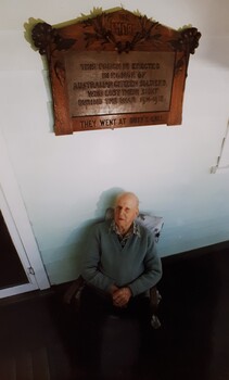 Elderly man sits beneath wooden plaque on verandah.