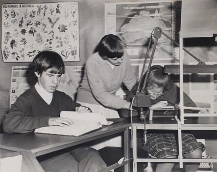 Schoolboy reading Braille and a schoolgirl assisted by a female teacher to read using lighting and other aids