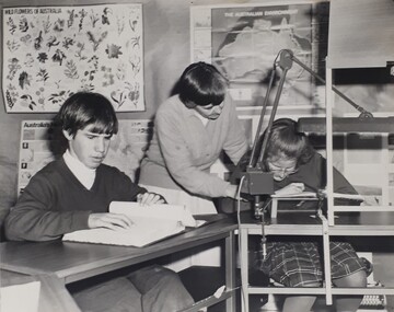 Schoolboy reading Braille and a schoolgirl assisted by a female teacher to read using lighting and other aids