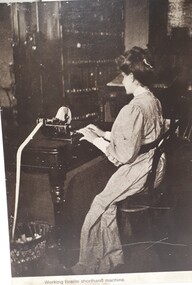 Woman in neck to floor dress, using a Braille shorthand machine in a room filled with books.