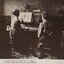 Girl plays an upright piano, reading the Braille score, under the supervision of a seated female teacher