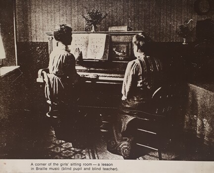 Girl plays an upright piano, reading the Braille score, under the supervision of a seated female teacher