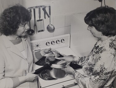 Two woman stand in front of a cooker, touching dials on a pressure cooker.