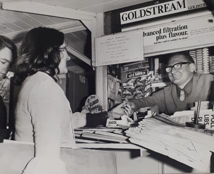 Woman buys from a newsagent at train station. Above them is a typewritten sign asking for help for Carols by Candlelight