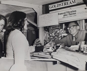 Woman buys from a newsagent at train station. Above them is a typewritten sign asking for help for Carols by Candlelight