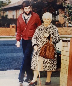 A bearded man walks next to an elderly woman holding a white cane