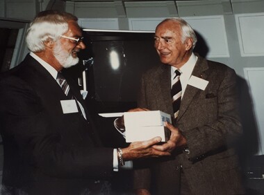 Silver haired man smiles as he presents two boxes of tapes to another silver haired man