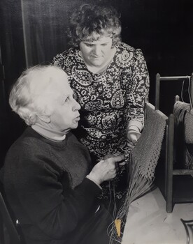 Elderly woman holds the threads, whilst another woman looks at the weaving the other has done.